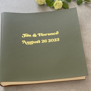 Peut inclure: Album photo vert olive fermé, avec lettrage doré. Le texte sur la couverture indique « Jim & Florence August 26 2023 ». L'album a un dos arrondi et un intérieur de couleur claire.
