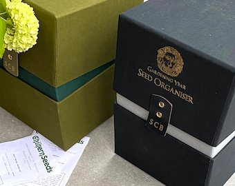 May include: Two boxes for organising seeds. The larger box is black with gold lettering that reads "Gardening Year Seed Organiser" and a gold lion emblem. The smaller box is green with a dark green stripe and a gold label that reads "SCB".