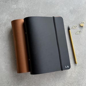 May include: Two notebooks, one brown and one black, are displayed on a gray surface. The black notebook has a black elastic band and the initials "L.B." in white. A gold pencil and paper clips are next to the notebooks.