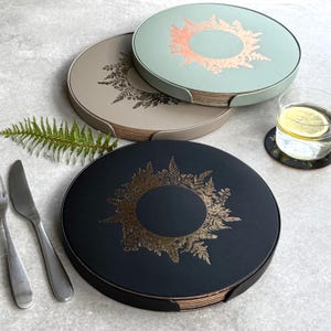 Könnte beinhalten: Runde Untersetzer-Sets in Schwarz, Beige und Mintgrün, jeweils mit goldenem Blumendesign. Ein Glas Wasser mit Zitrone und Besteck sind ebenfalls zu sehen. Durchmesser ca. 10 cm.