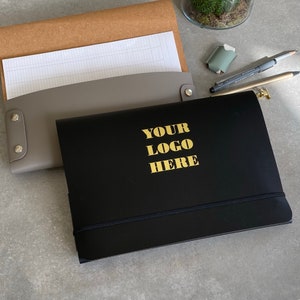 May include: A black notebook with the text "YOUR LOGO HERE" in gold lettering. A gray leather document holder and a brown paper folder are visible in the background. A pencil, pen, and eraser are also present.