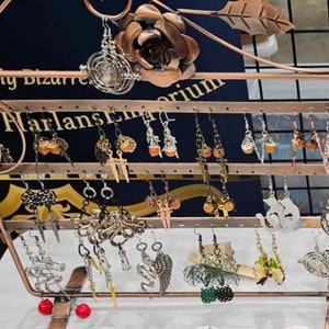 以下が含まれることがあります： イヤリングをディスプレイするための、複数のフックが付いた銅色の金属製スタンド。スタンドの上部には装飾的なバラのデザインが施されており、シルバー、ゴールド、ブラックなど、さまざまなデザインのイヤリングが飾られています。