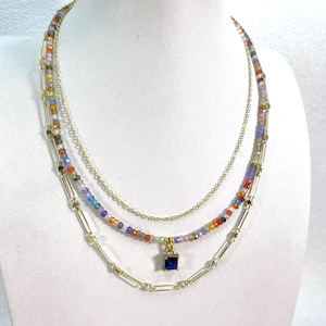 May include: A three-strand necklace displayed on a white mannequin. The top strand is a delicate gold chain. The middle strand features colorful beads. The bottom strand has gold bars and a square blue pendant.