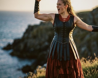 May include: A woman in a black leather bodice and a long, flowing red skirt stands on a cliff overlooking the ocean. She raises her fist in a gesture of triumph, wearing arm cuffs. The scene is bathed in warm sunlight.