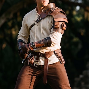 May include: Brown leather shoulder armor with small pouches and metal accents. The armor is paired with matching brown leather arm guards, gloves, and a belt with a pouch. The outfit is completed with a cream-colored shirt and brown pants.