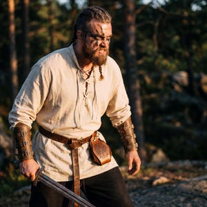 May include: A Viking-themed ensemble featuring a cream-colored tunic with a laced neckline, a brown leather belt, and matching arm bracers. The outfit includes a leather pouch and a sword. The person has face paint and braided hair.