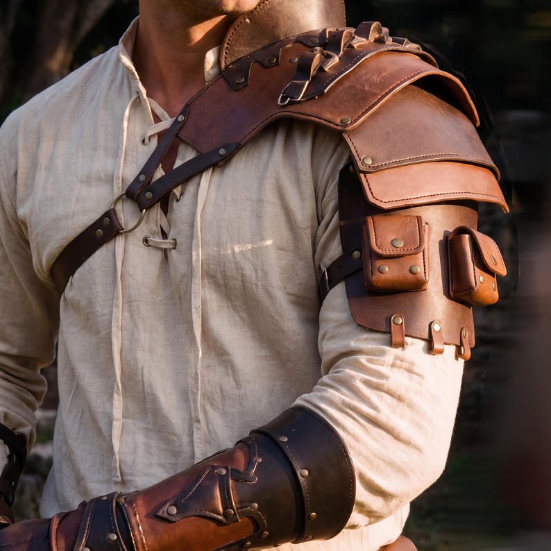May include: A brown leather shoulder armor piece with two small pouches and a matching arm guard. The armor is detailed with rivets and straps, and is worn over a light-colored long-sleeved shirt. The design is reminiscent of medieval or fantasy attire.