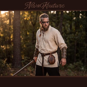 May include: A person in a cream-colored tunic with a laced neckline, leather belt, and arm cuffs, holding a wooden staff. A leather pouch with a tree design hangs from the belt. The image has the text "Nova Kouture" in the upper right corner.