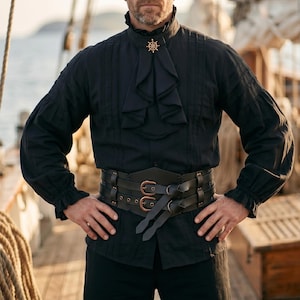 May include: Black pirate-style shirt with a ruffled collar and cuffs. The shirt features vertical pleats and a decorative gold-colored pin. A wide black leather belt with bronze buckles completes the look. The outfit is suitable for cosplay or themed events.