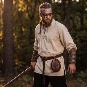 May include: A person in a cream-colored tunic and black pants, with a brown leather belt and pouch. The person is holding a wooden sword. The person has a beard, braided hair, and face paint, with leather arm guards.