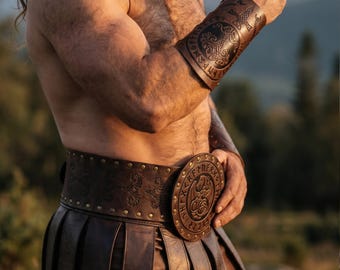 May include: Brown leather wrist cuff and belt with embossed designs. The cuff features a circular emblem with intricate patterns and runes. The belt has a large, round buckle with a similar design. The items appear to be part of a historical or fantasy costume.