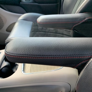 May include: Black leather car seat with red stitching and a beige center console. The seat has a built-in armrest with red stitching.