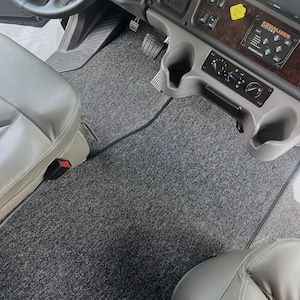 May include: Gray carpeted floor mats with black edging, installed in a vehicle. The mats cover the driver and passenger footwells. The driver's side mat has a red safety button.
