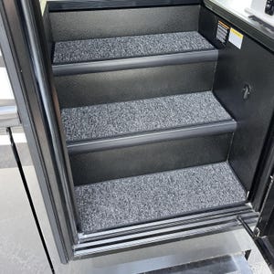 May include: A set of three black steps with gray speckled carpet treads. The steps are framed by a black metal structure, with a door frame visible on the right. The steps lead upwards, suggesting an entry point.