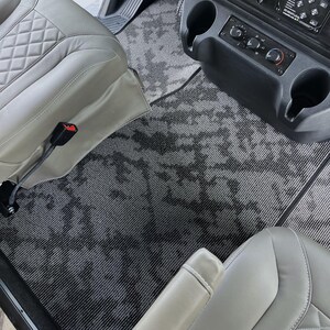 May include: Interior view of a vehicle featuring gray patterned floor mats and light gray leather seats. The mats have a dark gray abstract design. The seats have a diamond-stitched pattern. Cup holders and control panel are visible.