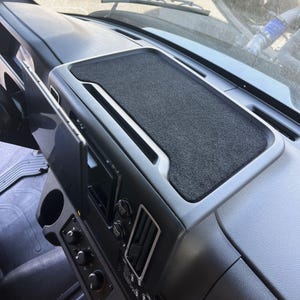 May include: A dark gray dashboard tray with a textured, felt-like surface. The tray is built into the dashboard of a vehicle, with a silver-colored trim. The dashboard also features air vents, control knobs, and a cup holder.