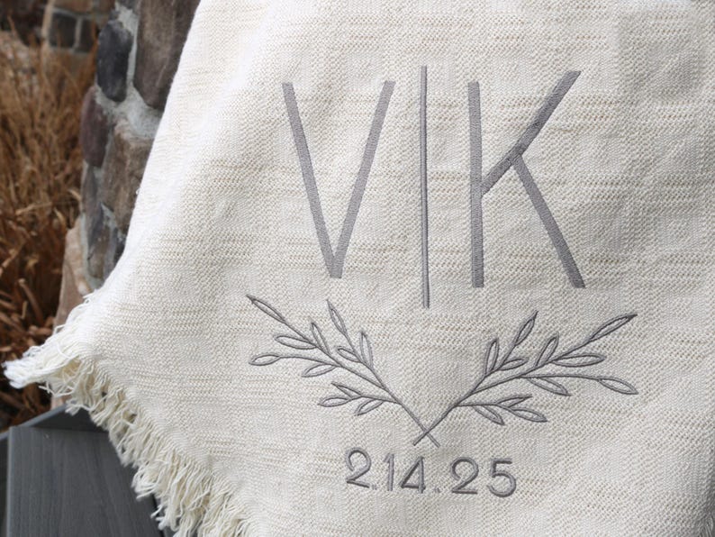 Peut inclure: Une couverture blanche avec un monogramme brod&eacute; gris "VK" et une date "2.14.25" sous une paire de branches crois&eacute;es.
