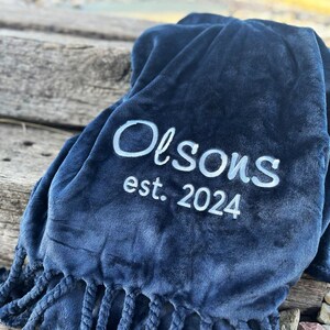 Peut inclure: Une couverture bleu marine avec le texte brod&eacute; blanc "Olson's est. 2024". La couverture a une texture douce et moelleuse et des franges d&eacute;coratives sur le bord inf&eacute;rieur. La couverture est pli&eacute;e et drap&eacute;e.