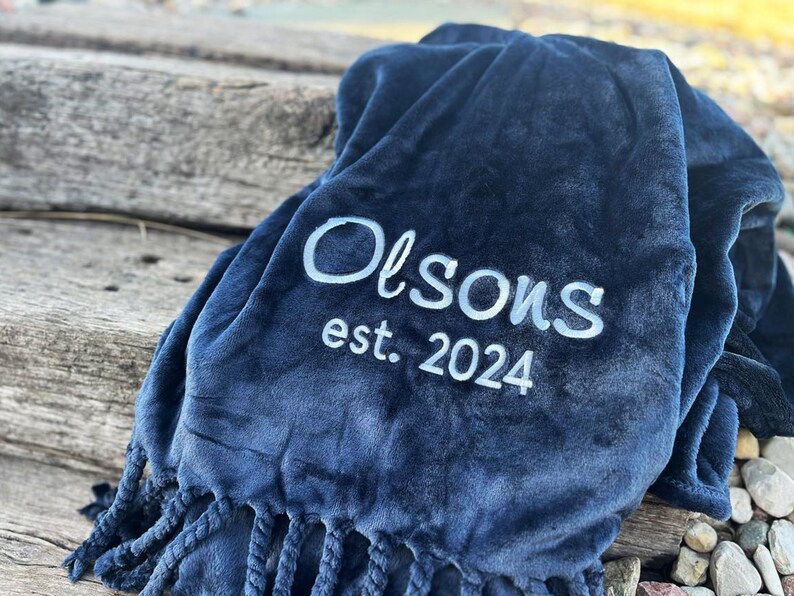 Peut inclure: Une couverture bleu marine avec le texte brod&eacute; blanc "Olson's est. 2024". La couverture a une texture douce et moelleuse et des franges d&eacute;coratives sur le bord inf&eacute;rieur. La couverture est pli&eacute;e et drap&eacute;e.