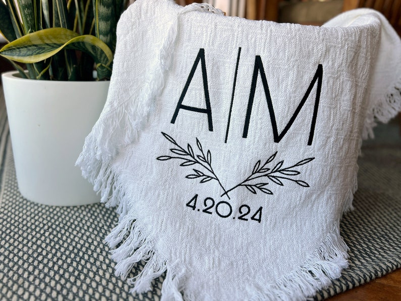 Pu&ograve; includere: Asciugamano da t&egrave; in cotone bianco con bordi sfrangiati, con iniziali ricamate in nero "A/M" e un motivo floreale con la data "4.20.24" sotto.
