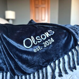 Peut inclure: Une couverture en peluche bleu marine avec le texte brod&eacute; blanc "Olson's est. 2024." La couverture a un bord &agrave; franges et est drap&eacute;e sur un lit. La texture douce et velout&eacute;e est visible.