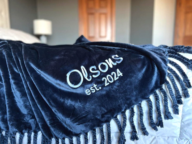Peut inclure: Une couverture en peluche bleu marine avec le texte brod&eacute; blanc "Olson's est. 2024." La couverture a un bord &agrave; franges et est drap&eacute;e sur un lit. La texture douce et velout&eacute;e est visible.