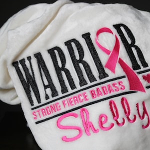 May include: A white, plush blanket embroidered with the word "WARRIOR" in black, a pink ribbon, and the words "STRONG FIERCE BADASS". The name "Shelly" is embroidered in pink script below. A heart is embroidered next to the ribbon.