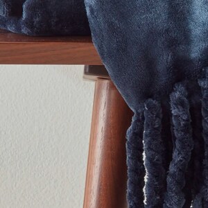 Peut inclure: Une couverture en peluche bleu marine avec une bordure &agrave; franges. La couverture est drap&eacute;e sur une table en bois fonc&eacute;, mettant en valeur sa texture douce et sa couleur profonde. La frange ajoute un &eacute;l&eacute;ment d&eacute;coratif.