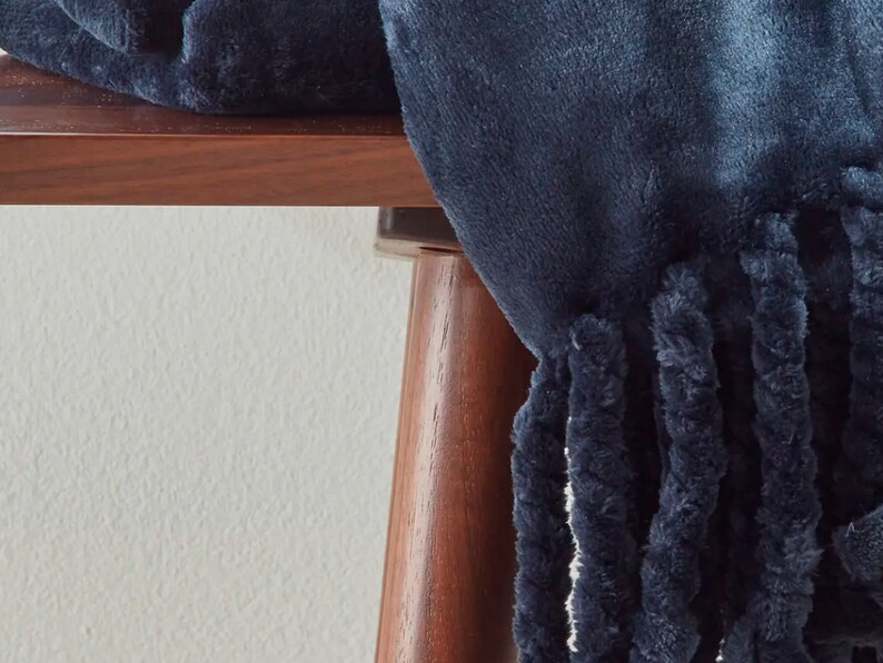 Peut inclure: Une couverture en peluche bleu marine avec une bordure &agrave; franges. La couverture est drap&eacute;e sur une table en bois fonc&eacute;, mettant en valeur sa texture douce et sa couleur profonde. La frange ajoute un &eacute;l&eacute;ment d&eacute;coratif.