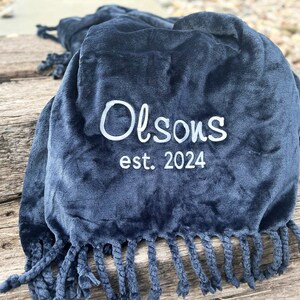 Peut inclure: Une couverture en peluche bleu marine avec une bordure &agrave; franges. Le texte brod&eacute; "Olson's est. 2024" est en blanc. La couverture est pli&eacute;e, mettant en valeur sa texture douce et son design.