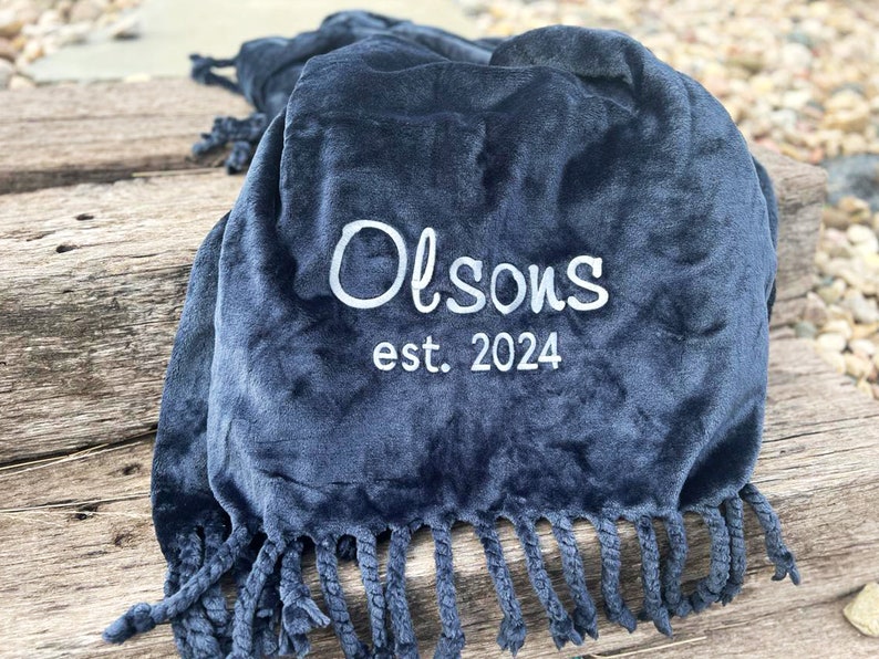 Peut inclure: Une couverture en peluche bleu marine avec une bordure &agrave; franges. Le texte brod&eacute; "Olson's est. 2024" est en blanc. La couverture est pli&eacute;e, mettant en valeur sa texture douce et son design.
