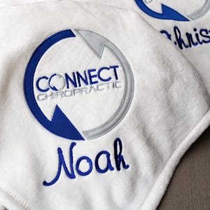 Puede incluir: Mantas de forro polar blancas bordadas con el logotipo "Connect Chiropractic" en azul y gris. Una manta está personalizada con el nombre "Noah" en escritura azul, y otra con "Christine". Las mantas son suaves y esponjosas.