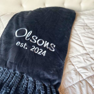 Peut inclure: Une couverture bleu marine avec une texture douce et velout&eacute;e et une bordure &agrave; franges. La couverture est brod&eacute;e du nom "Olson" et de l'ann&eacute;e "est. 2024" en police blanche. La couverture est pos&eacute;e sur un lit.