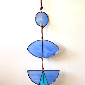 Puede incluir: Un móvil de vidrio tintado con tres piezas de vidrio azul que cuelgan de un cordón de cuero marrón. La pieza superior es un círculo, la pieza del medio es un óvalo y la pieza inferior tiene forma de media luna.