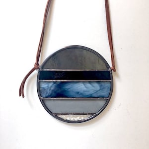 Könnte beinhalten: Ein runder Buntglas-Sonnenfänger mit einem braunen Lederband. Der Sonnenfänger hat fünf horizontale Glasbänder in Grautönen und Blau.