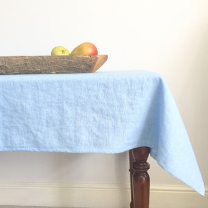 Op de afbeelding: Een lichtblauw linnen tafelkleed gedrapeerd over een houten tafel met een houten kom met twee appels.