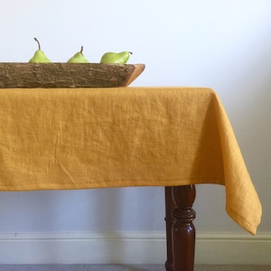 May include: A mustard yellow linen tablecloth draped over a wooden table with three green pears in a wooden bowl.