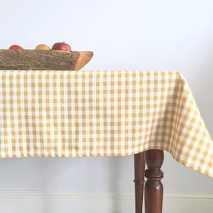 May include: A yellow and white gingham tablecloth draped over a wooden table with a wooden bowl filled with apples on top.