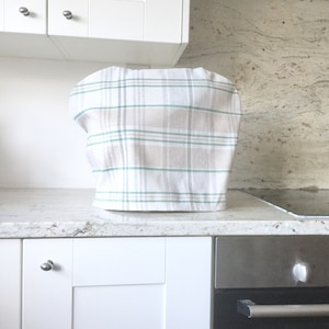 May include: A white kitchen counter with a white and green plaid kitchen appliance cover. The counter has a white cabinet below it and a stainless steel oven with a black door below that.