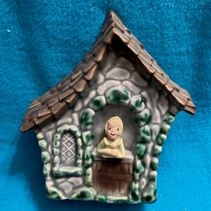 May include: A ceramic cottage wall decor with a green roof and a small figure of a woman with yellow hair and a green dress looking out the window.