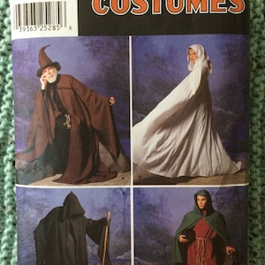 May include: A Simplicity Costumes sewing pattern envelope, number 9887, featuring four variations of hooded cloaks and robes in brown, white, black, and green. The packaging includes a barcode, size range XS to XL, and the brand name Simplicity.