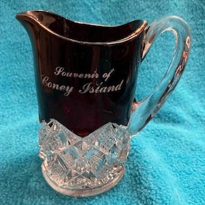 May include: A dark purple glass pitcher with a clear glass handle. The pitcher has a diamond-shaped pattern on the bottom and says "Souvenir of Coney Island" on the front.