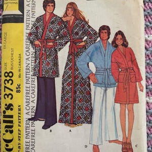 May include: Vintage McCall's 3738 sewing pattern for robes. The pattern features illustrations of robes in various styles, including a long patterned robe, a short red robe, and a blue robe with white pants. The pattern is labeled "Extra Carefree".