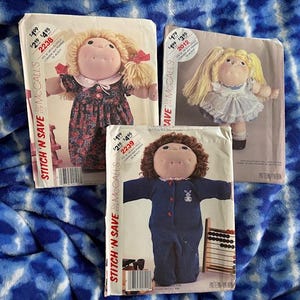 May include: Three vintage McCall's Stitch 'N Save sewing patterns for dolls. The patterns feature dolls in various outfits, including a floral dress, a white dress, and a blue jumpsuit. The pattern numbers are 2238, 2239, and 2012.