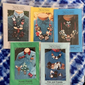 May include: Five different sewing patterns for felt necklaces. Each pattern features a different theme: Forest Friends, Fido and Friends, Home Friends, and two others with a moose and a girl. The patterns are printed on white paper with a blue and white floral background.