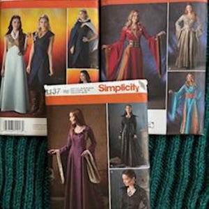 May include: Three Simplicity sewing pattern envelopes labeled 1246, 1487, and 1137. Each envelope displays photographs of long, flowing dresses and capes in various colors including red, purple, blue, and white, with detailed garment construction illustrations.