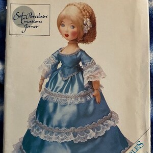May include: A Butterick craft pattern featuring a porcelain doll in a blue dress with white lace trim. The doll has blonde hair and is wearing a blue dress with white lace trim. The pattern is labeled "Soft Porcelain Creations Janet" and "Collectibles 1860".