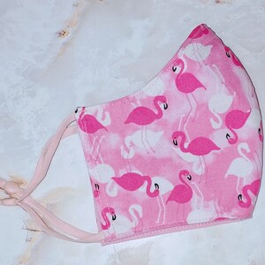 May include: Pink face mask with a white background and a flamingo print. The mask has two pink ties.