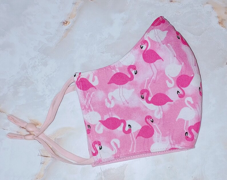 May include: Pink face mask with a white background and a flamingo print. The mask has two pink ties.