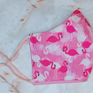 May include: Pink face mask with a flamingo print. The mask has adjustable ear loops and a pink and white pattern.